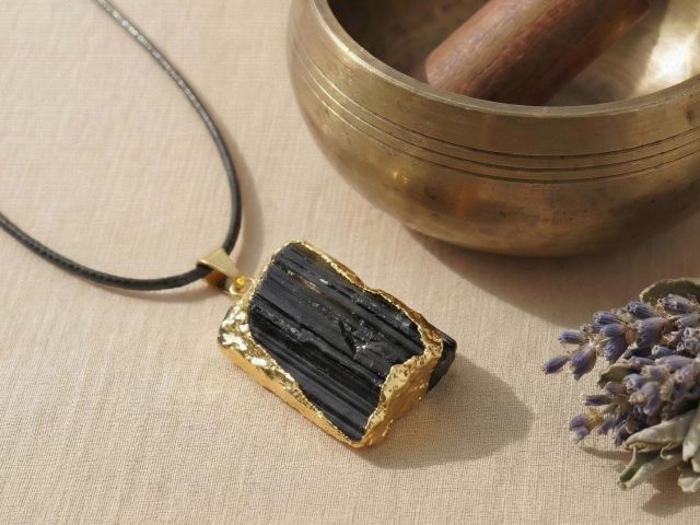 Nahaufnahme eines schwarzen Turmalin-Anhängers mit Goldrand auf beigem Stoff neben einer Klangschale.