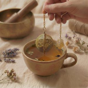 Ein goldenes Teesieb hängt an einer Kette mit Citrin-Stein über einer hellen Keramiktasse.