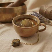 Goldenes Teesieb schwimmt in einer dampfenden Tasse Tee, derTigerauge Chakren Stein liegt dekorativ daneben.