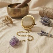 Hand hält das goldene Tee-Ei am Amethyst-Anhänger über eine beige Keramiktasse auf Leinenstoff.