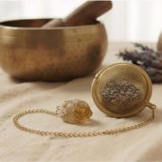 Hand hält das goldene Tee-Ei am funkelnden Citrin-Anhänger über eine weiße Schale.