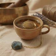 Ein goldenes, rundes Teesieb wird an einer Kette über eine rustikale, dunkle Keramiktasse gehalten, wobei der Labradorit-Stein bläulich im Licht aufleuchtet.