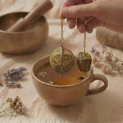 Ein goldenes, rundes Teesieb mit Tigerauge Edelstein wird an einer Kette über eine handgetöpferte Keramikschale mit Wasser gehalten.