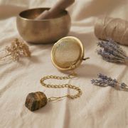 Hand hält das goldene Tee-Ei am Amethyst-Anhänger über eine beige Keramiktasse auf Leinenstoff.